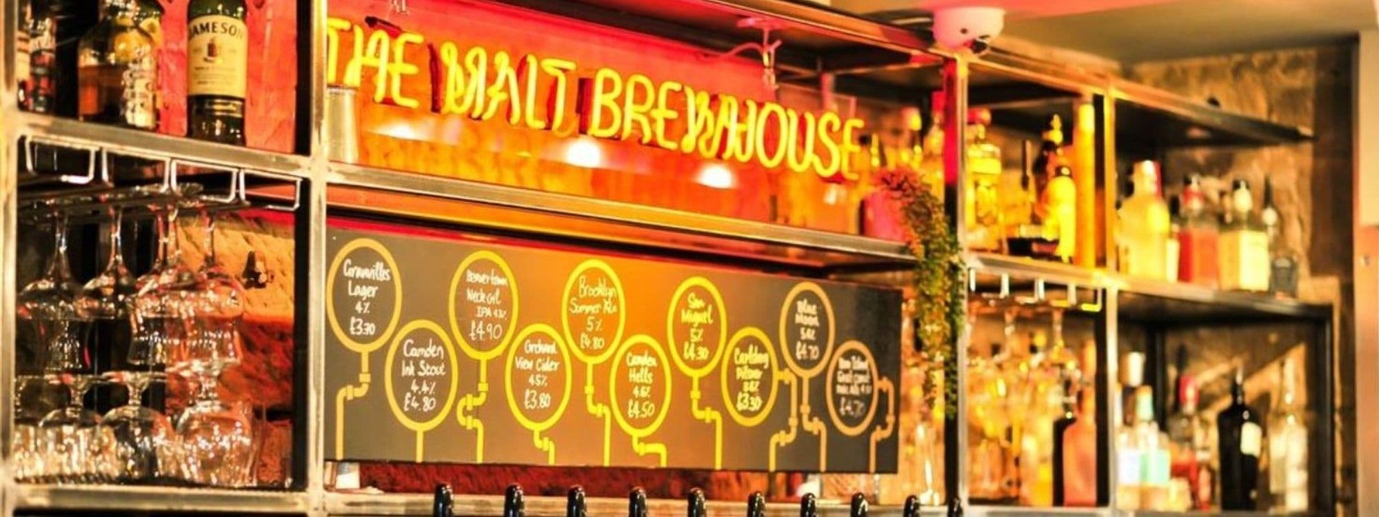 Interior view of The Malt Brewhouse featuring a vibrant neon sign, a variety of beers on tap with prices listed on illuminated round boards, and a selection of spirits and glasses on shelves.