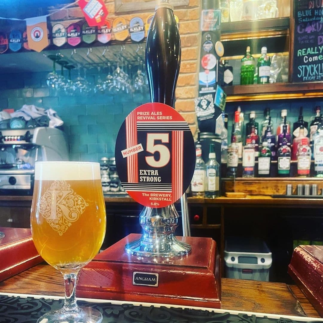 A pint of beer in a branded glass on a tray, with a beer tap featuring a "Number 5 Extra Strong" ale badge in the foreground at The Old Kings Arms. Behind, a bar with various spirit bottles and a display of beer tap badges.