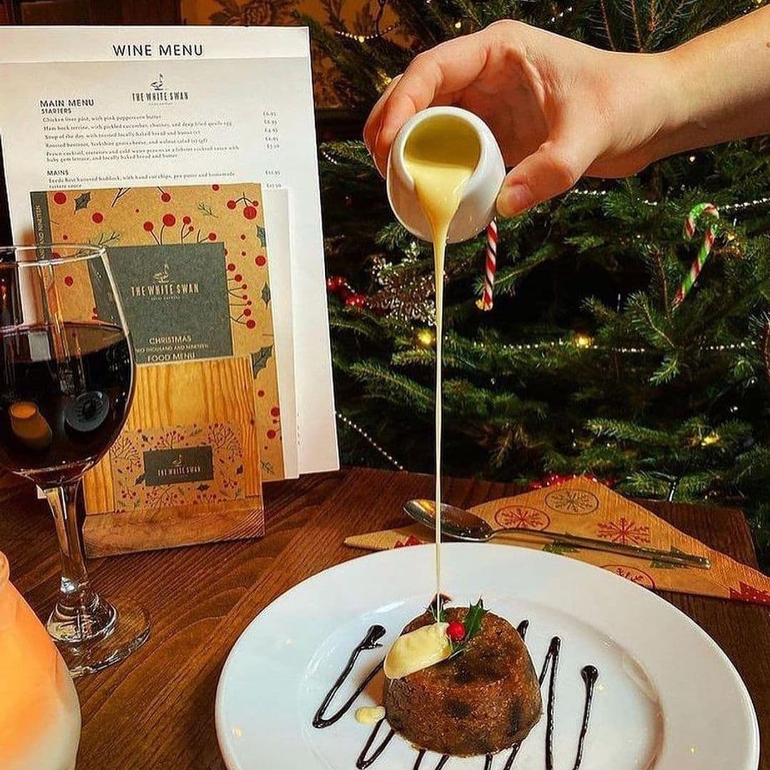 Alt text: "Enjoy a festive dining experience at The White Swan with a traditional Christmas pudding being drizzled with creamy custard, accompanied by a glass of red wine and a holiday-themed menu in the background, all set against the cozy ambiance of a decorated Christmas tree."
