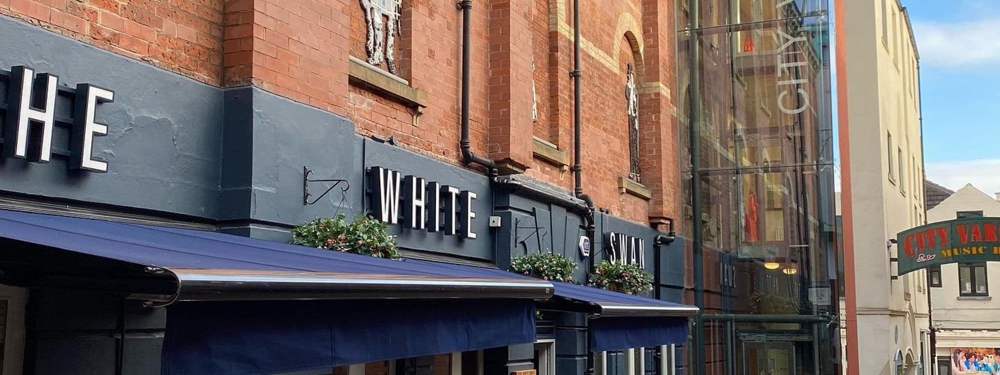 Exterior view of The White Swan, showcasing the establishment's bold signage above blue awnings adorned with greenery, set against a backdrop of traditional brick buildings.