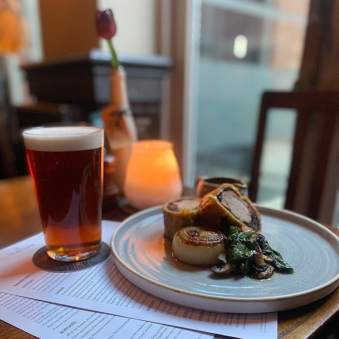 Gourmet dining experience at The White Swan featuring a pint of amber ale, seared scallops, and a plate of rolled meat with spinach and mushrooms, set on a table with a menu.