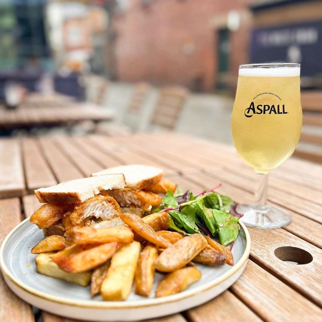 Alt text: "Enjoy a casual dining experience at The White Swan with a plate of fish and chips accompanied by a fresh salad and a chilled glass of Aspall cider on a wooden outdoor table."