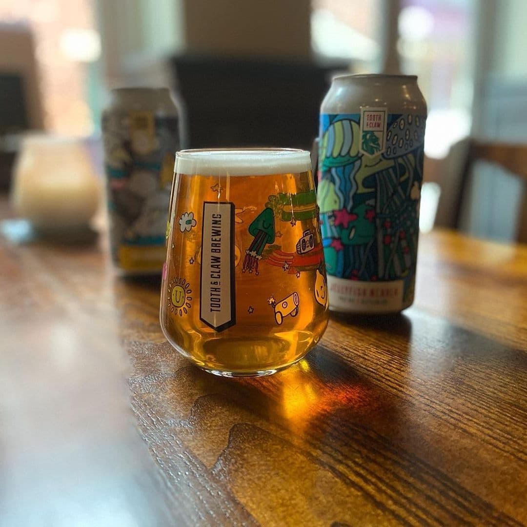A glass of beer on a wooden table with colorful illustrated cans in the background at The White Swan.