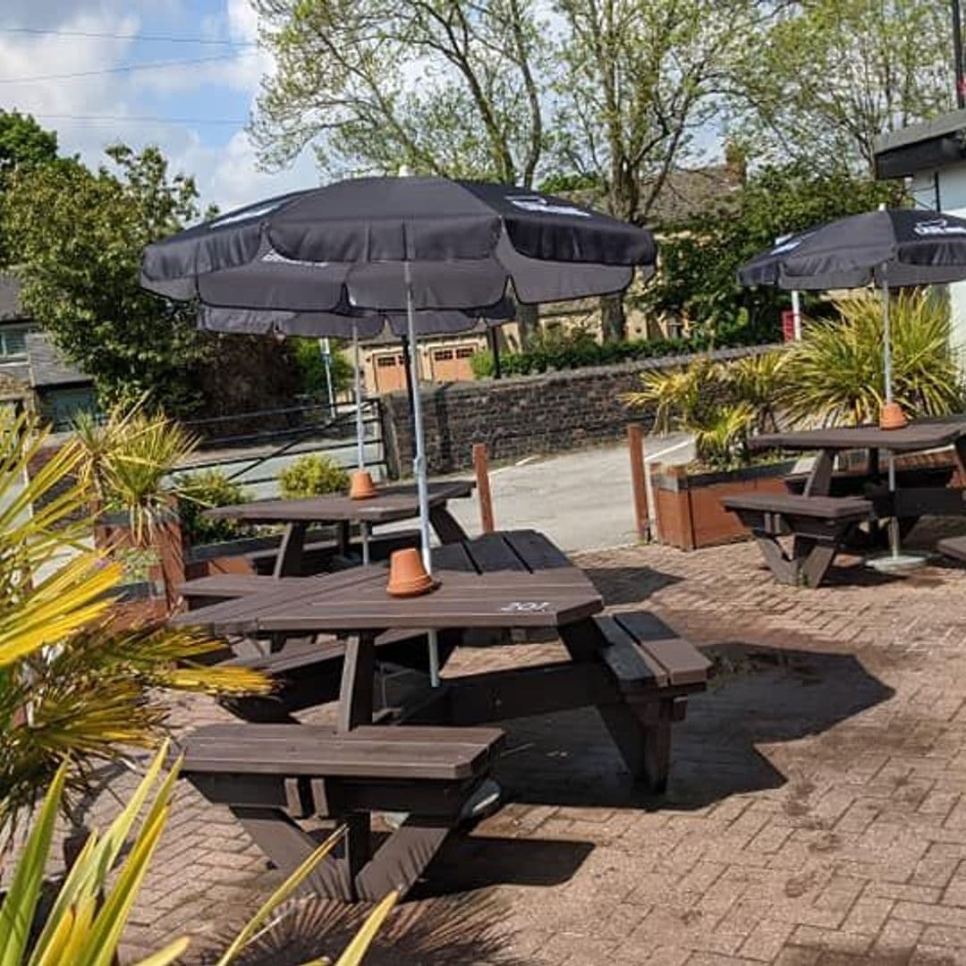 Outdoor seating area with wooden picnic tables and umbrellas at The Woodcock New Farnley, surrounded by greenery and a sunny sky.