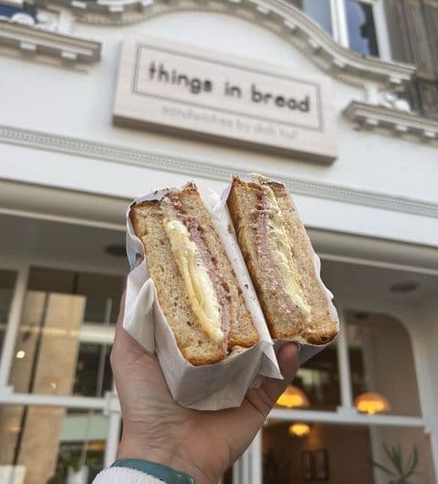 Hand holding a sandwich in front of "Things in Bread" sandwich shop with visible sign.