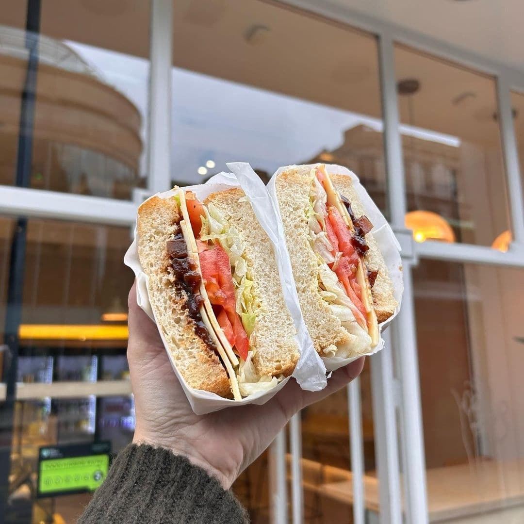 Hand holding a freshly made sandwich with lettuce, tomatoes, and sliced meat on whole grain bread in front of the Things in Bread shop.