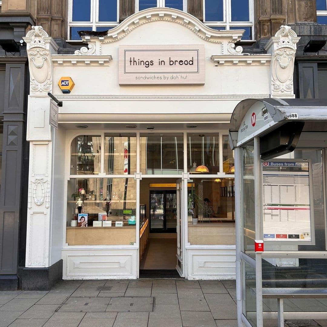 Exterior view of 'Things in Bread' sandwich shop with a classic facade, large windows, and an entrance flanked by white columns. A bus stop is visible in the foreground.