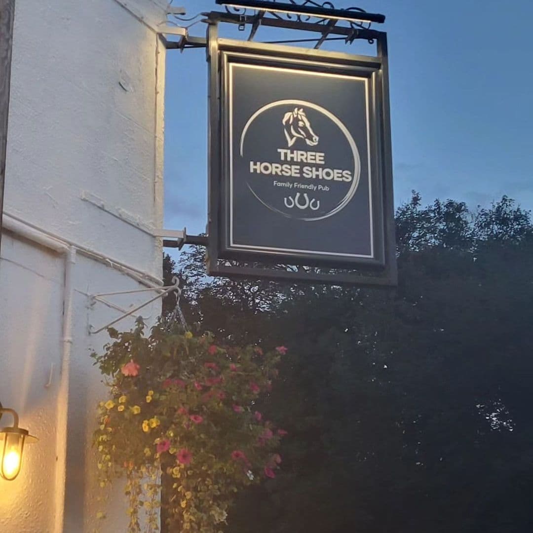 Sign of Three Horse Shoes Oulton, a family-friendly pub, with a blooming flower basket and a warm wall lantern at dusk.