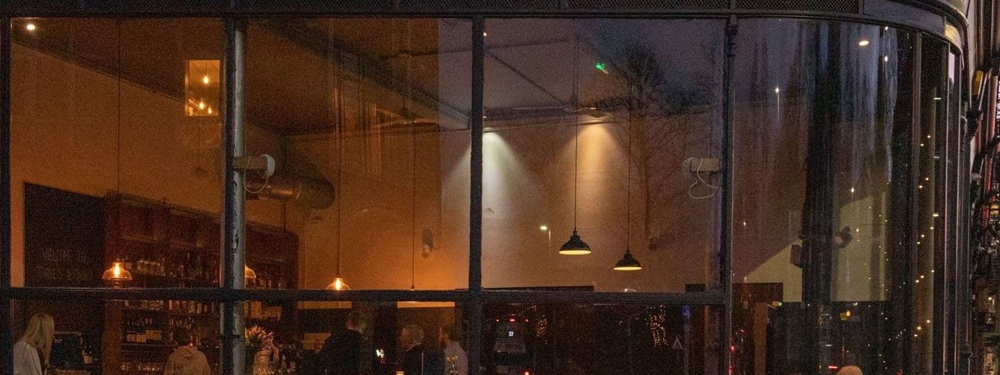 Evening view of the exterior of Three’s A Crowd, showcasing the warm interior lighting and patrons inside through the large glass windows.