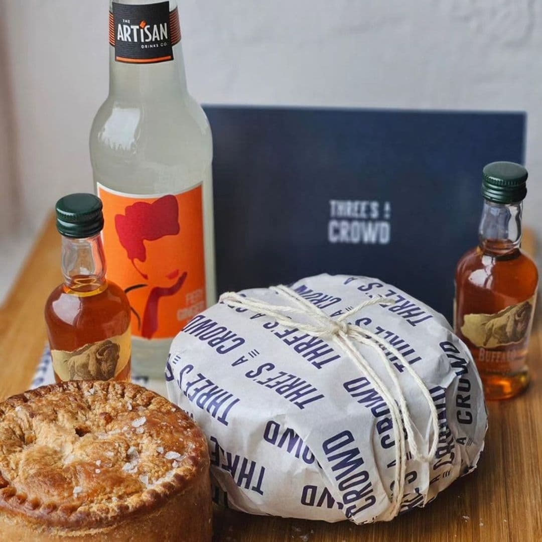 A selection of artisanal food and drink items from Three's A Crowd, featuring a wrapped pie, a bottle of craft soda, and two miniature bottles of spirits, with a branded menu in the background.