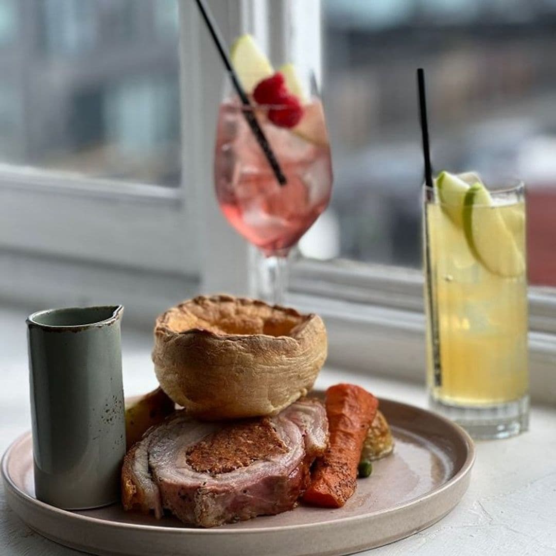 Gourmet meal at Three's A Crowd featuring roasted pork, Yorkshire pudding, carrots, and gravy, with refreshing cocktails by a window with a city view.