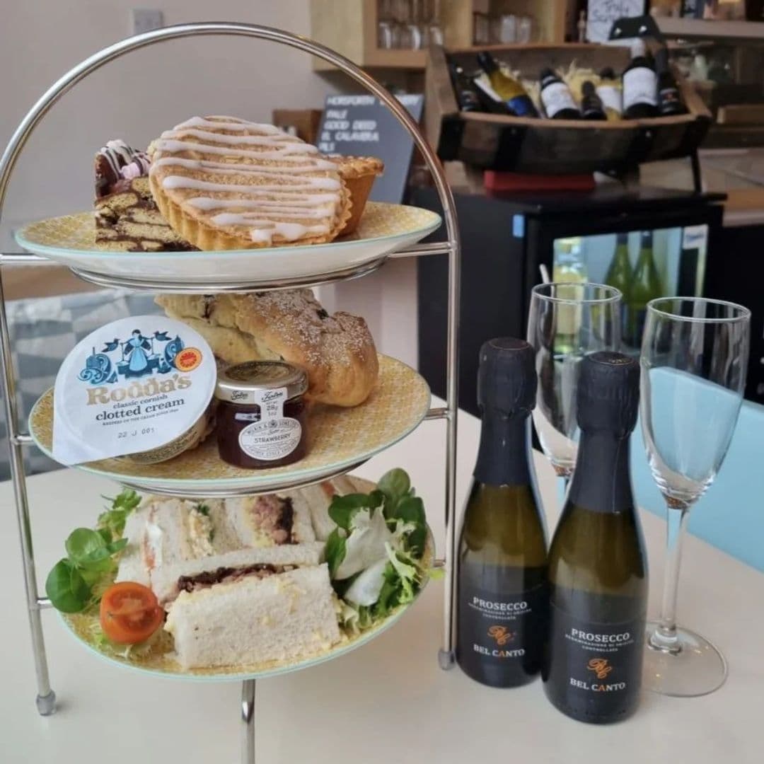 Afternoon tea setup at Truly Scrummy featuring a three-tiered serving stand with sandwiches, scones, clotted cream, and sweet pastries, accompanied by two bottles of Prosecco and filled glasses.