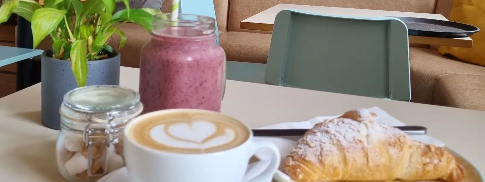 Alt text: "Enjoy a cozy breakfast at Truly Scrummy with a freshly brewed cappuccino with heart-shaped latte art, a berry smoothie in a glass jar with a straw, and a flaky, powdered sugar-dusted croissant on a white plate, complemented by a green potted plant and casual cafe seating in the background."