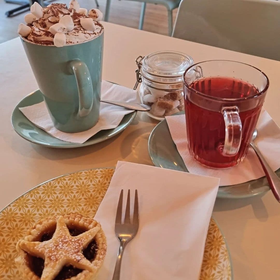 Alt text: "A cozy table setting at Truly Scrummy featuring a large mug of hot chocolate topped with whipped cream and marshmallows, a clear glass of red herbal tea, a mini star-topped pie, and a jar of brown sugar cubes, all served on a patterned tabletop."