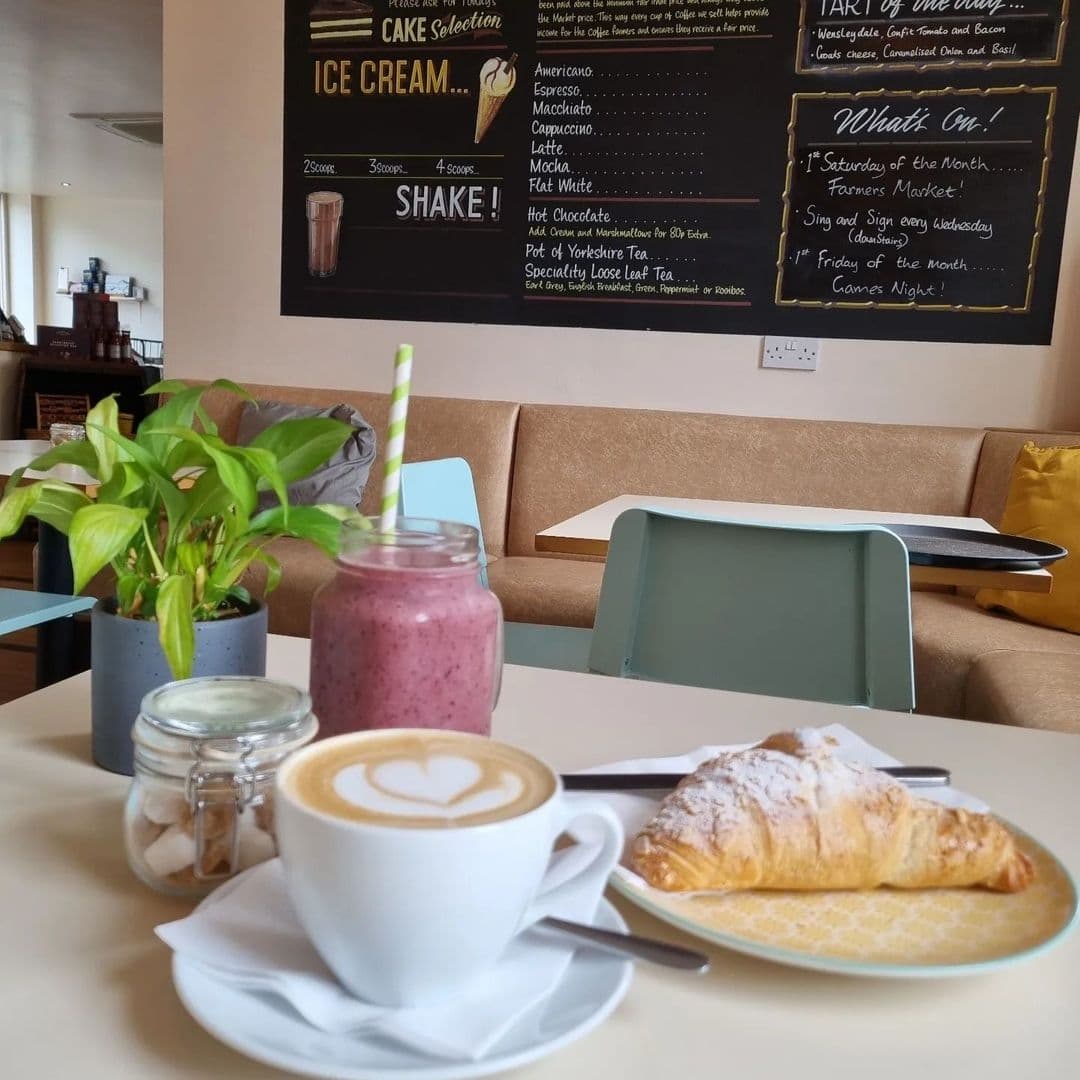 A cozy cafe setting at Truly Scrummy with a freshly brewed cappuccino with heart-shaped latte art, a berry smoothie in a mason jar with a striped straw, and a dusted croissant on a plate, all on a light-colored table with a potted plant and sugar cubes in a jar; a menu board in the background lists various beverages and events.