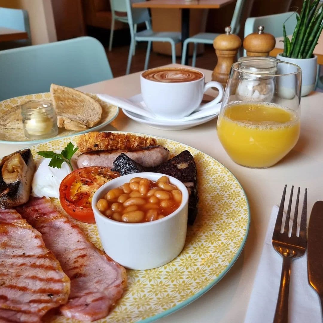 Alt text: "Hearty breakfast at Truly Scrummy featuring toast with butter, bacon, sausages, grilled tomato, black pudding, baked beans, mushrooms, a poached egg, a cup of coffee with latte art, and a glass of orange juice, all served on a floral patterned plate with cutlery on the side."