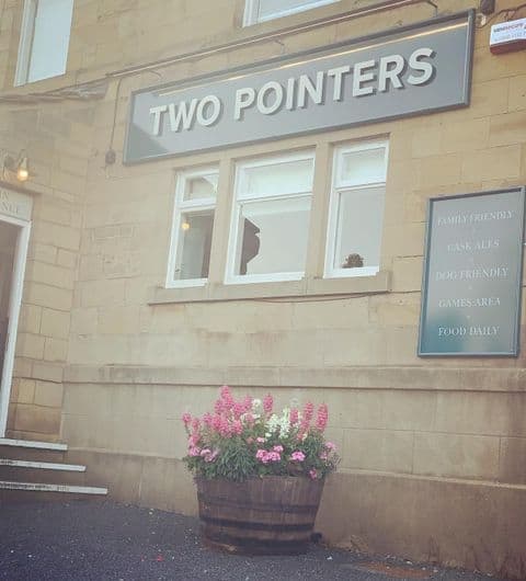 Exterior view of Two Pointers showing the establishment's sign, a broken window, and a signboard with text 'Family Friendly, Cask Ales, Dog Friendly, Games Area, Food Daily' next to a large flower pot with pink blooms.