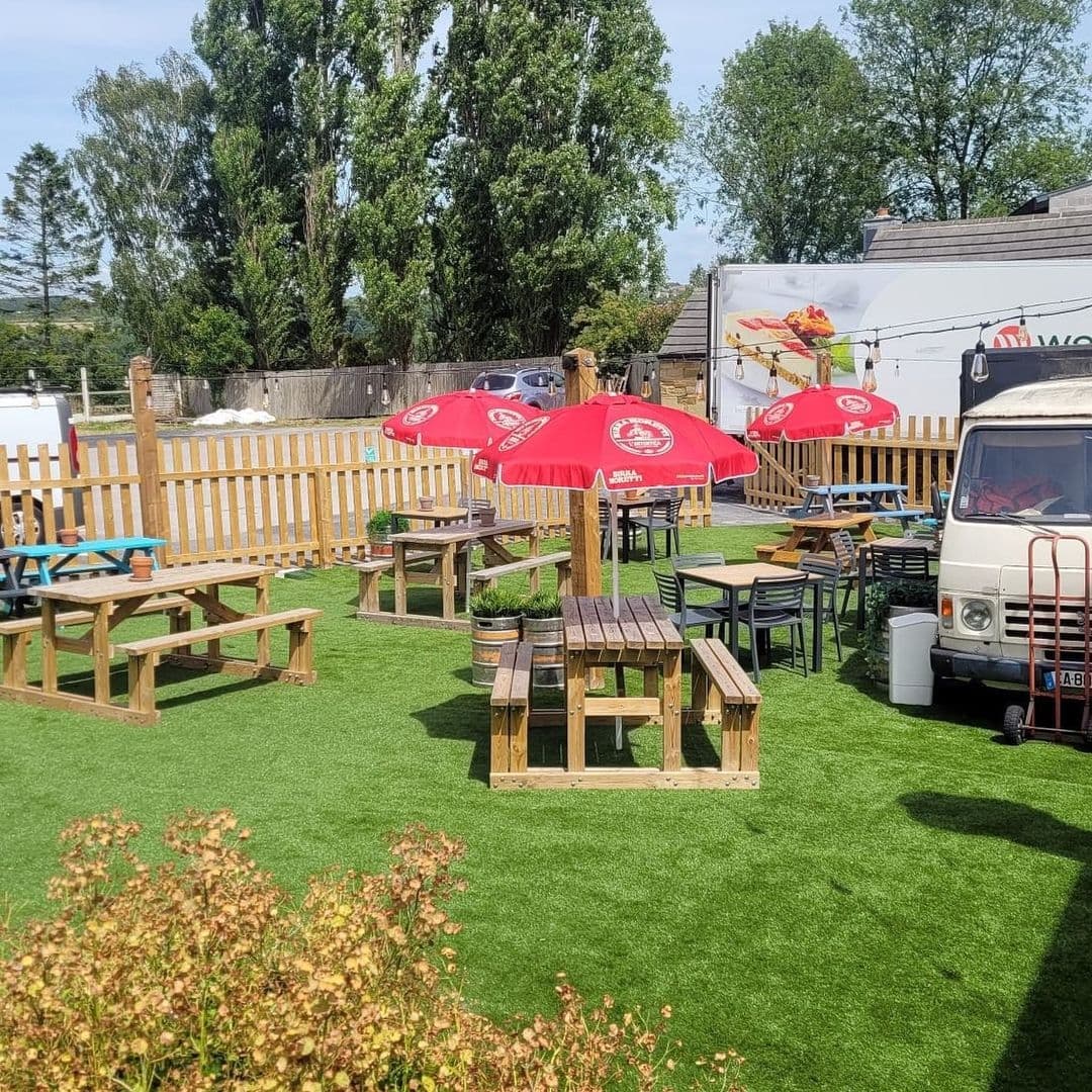 Sunny outdoor seating area at Two Pointers with wooden picnic tables, red umbrellas, artificial grass, and surrounding greenery.