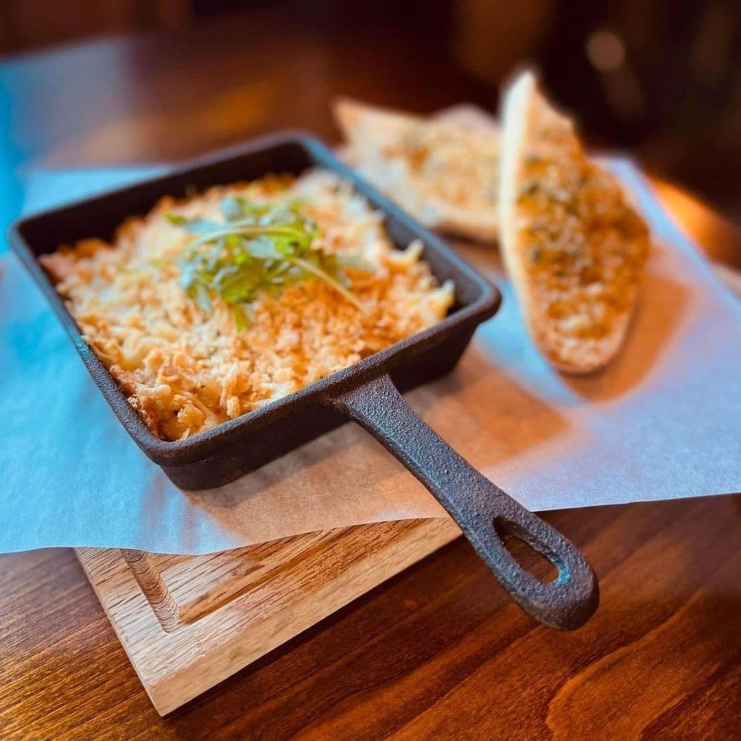 A freshly baked macaroni and cheese in a cast iron skillet, garnished with herbs, served with toasted bread on a wooden table at Two Pointers.
