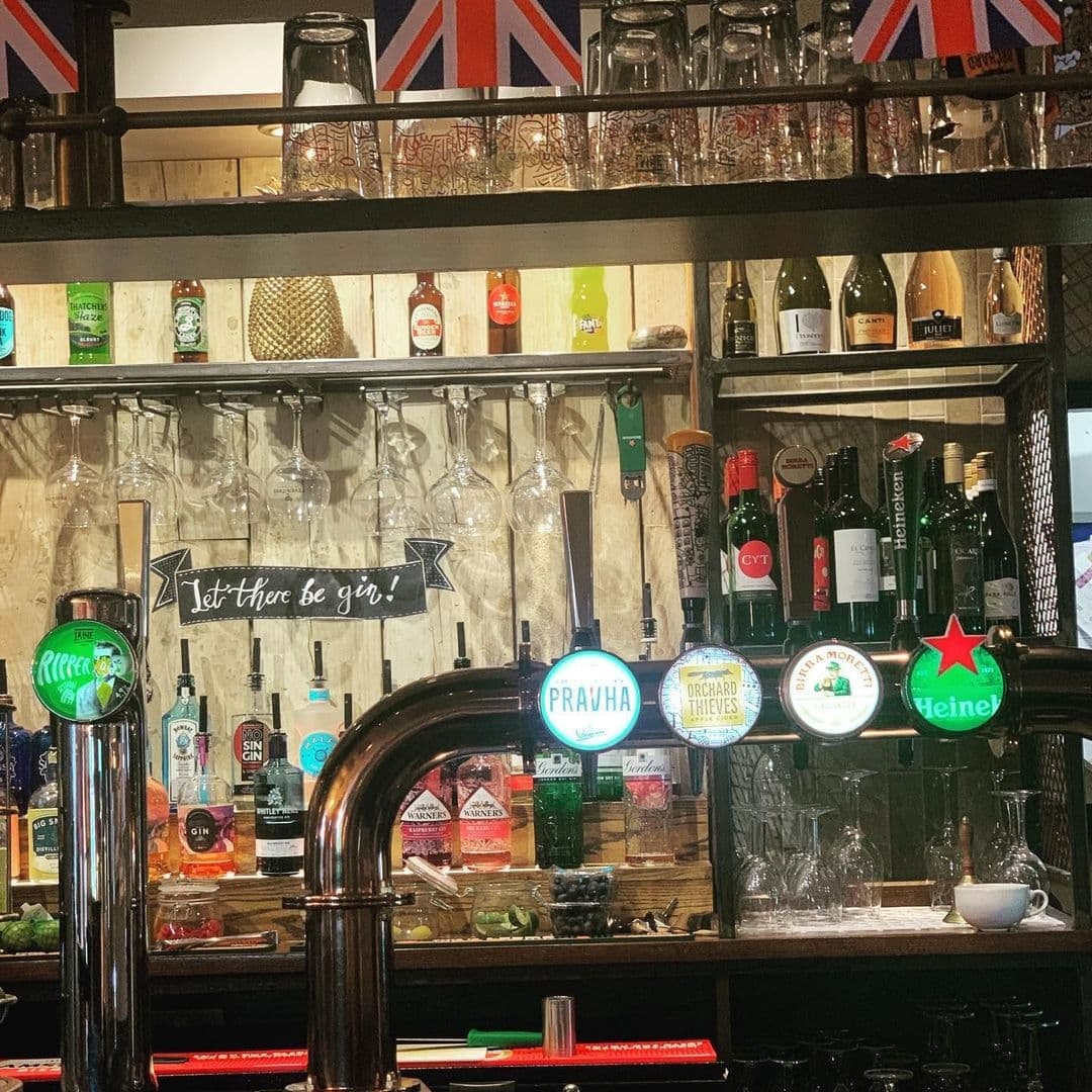Pub interior at Two Pointers featuring a variety of alcoholic beverages on shelves and taps, with a sign reading "Let there be gin!", and British flags above.