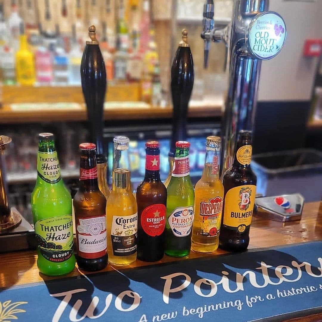 A variety of bottled beers displayed on a bar counter with beer taps in the background, featuring brands like Thatcher's, Budweiser, Corona, and others, at Two Pointers.