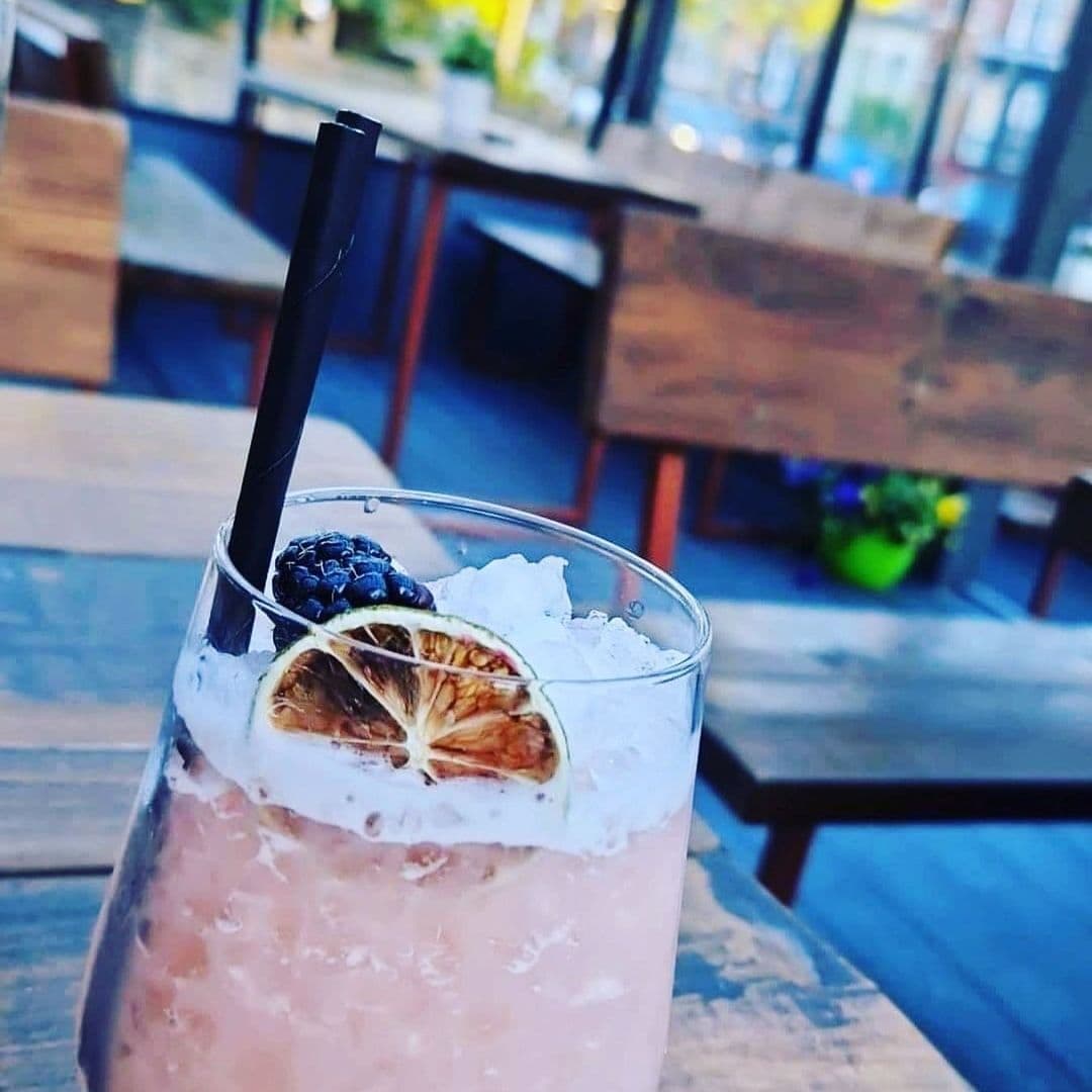 Close-up of a refreshing cocktail with a black straw, garnished with a blackberry and a dried citrus slice at Victoria Social, with blurred wooden tables and chairs in the background.