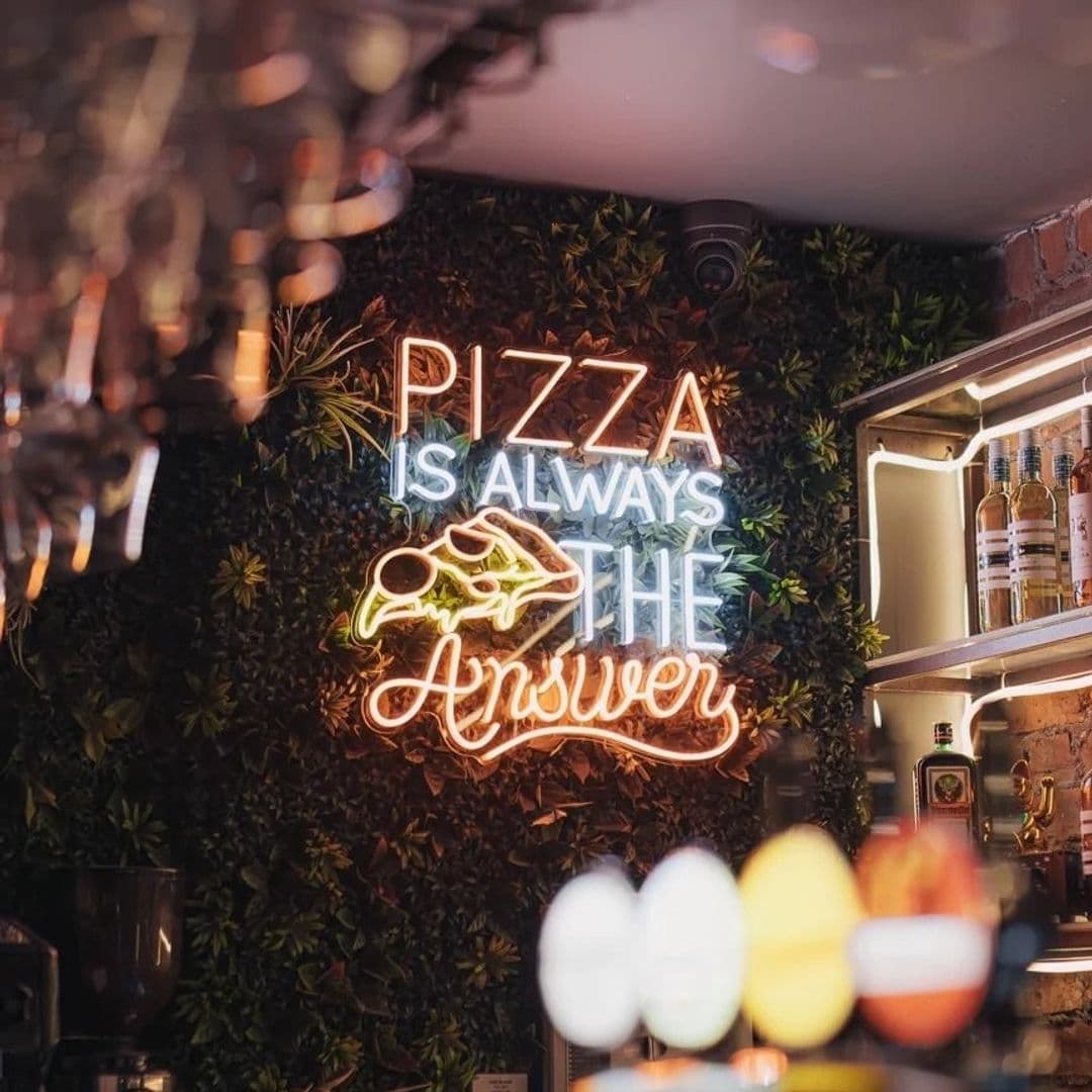 Neon sign stating "PIZZA IS ALWAYS THE ANSWER" on a leafy wall at Victoria Social, with blurred bar atmosphere in the foreground.