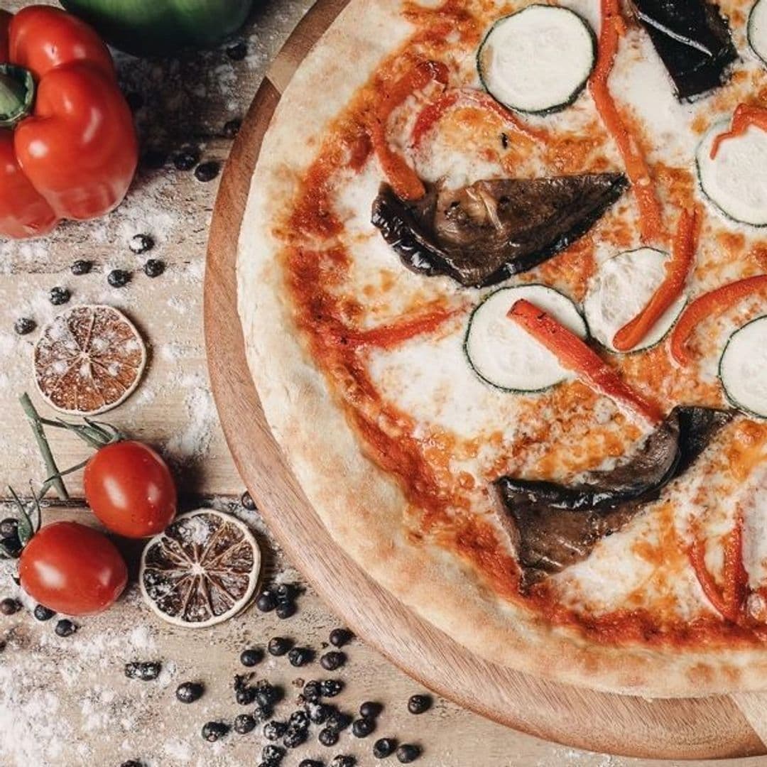 Freshly baked pizza with mozzarella, eggplant, zucchini, and bell pepper slices on a wooden serving board, surrounded by ingredients like tomatoes, black olives, and herbs at Victoria Social.
