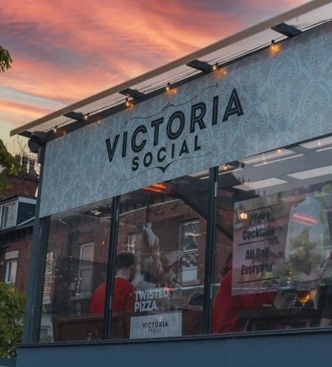 Exterior view of Victoria Social with illuminated signage, featuring a glass facade, patrons inside, and string lights under a twilight sky.