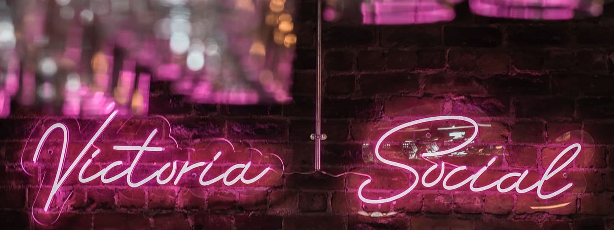 Neon sign reading 'Victoria Social' on a brick wall with a blurred pink light background.