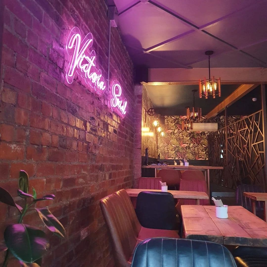 Cozy interior of Victoria Social with pink neon sign on exposed brick wall, plush seating, and ambient lighting.