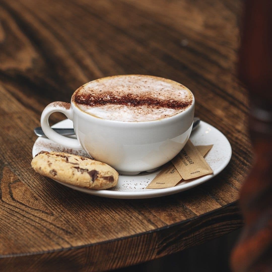 A freshly prepared cappuccino with a dusting of cocoa on top, accompanied by a chocolate chip biscuit, served on a wooden table at Woolpack Inn.