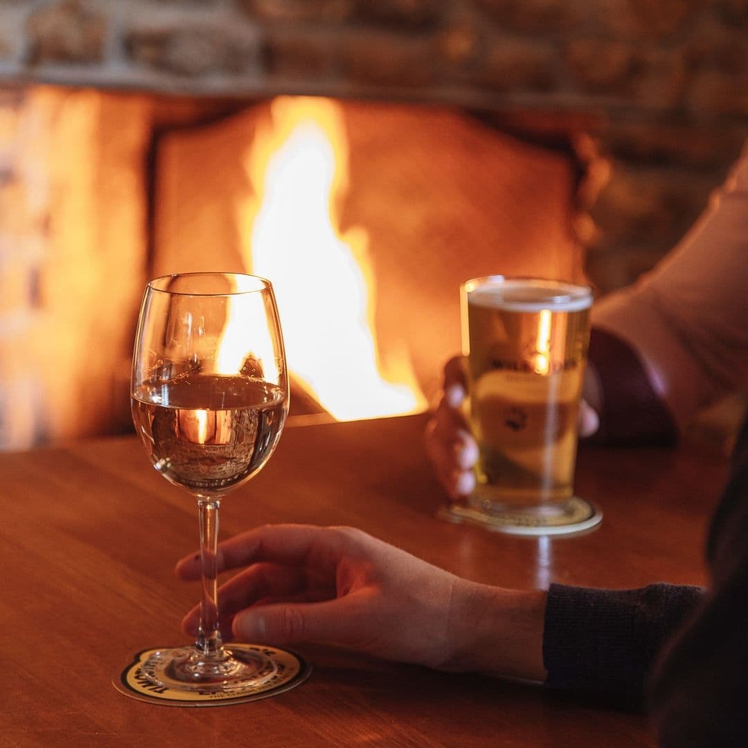Cozy atmosphere at Woolpack Inn with a close-up of a wine glass and a hand holding a pint of beer, both resting on a wooden table, with a warm fireplace in the background.