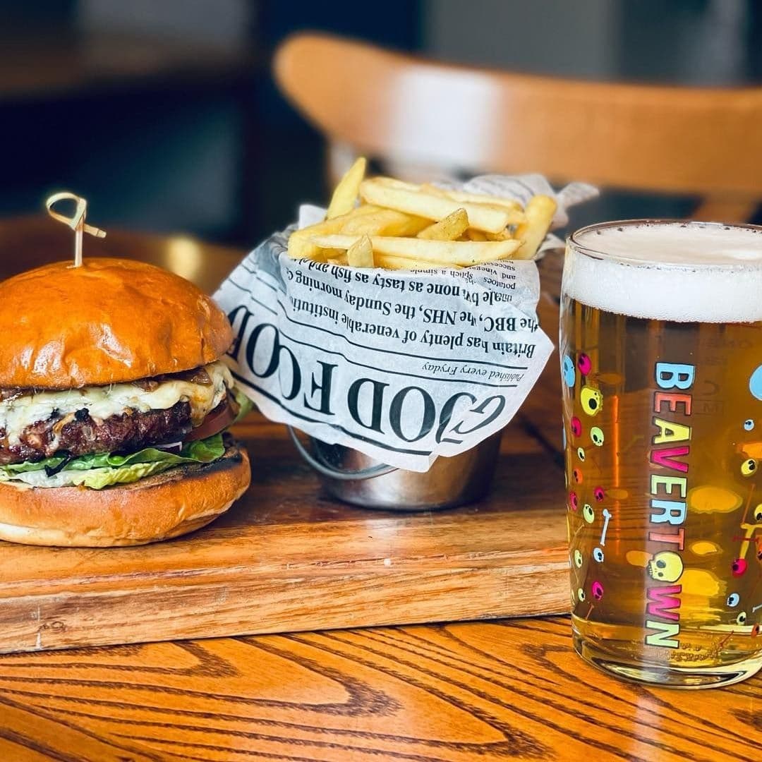 Juicy burger with lettuce and cheese on a wooden table at Woolpack Inn, accompanied by a pint of beer and a side of fries in a metal bucket with newspaper lining.