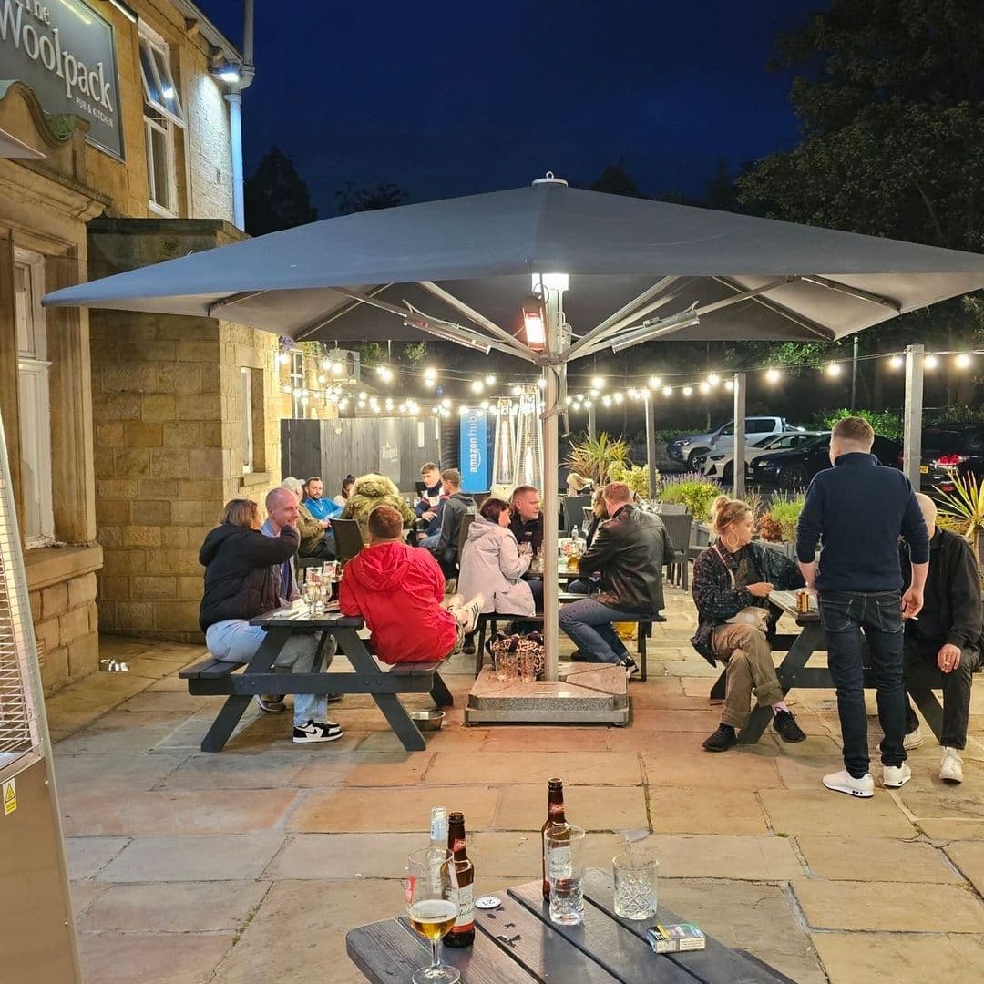 Outdoor evening setting at Woolpack Inn with guests enjoying drinks at picnic tables under a patio umbrella and string lights.