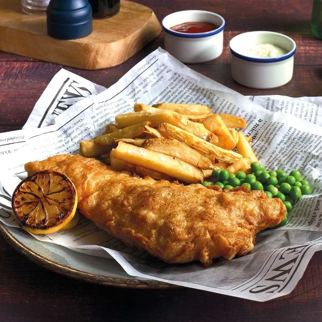 Traditional fish and chips served on a newspaper with peas and a grilled lemon slice at Woolpack Inn. Condiments in small bowls and a pepper grinder in the background.