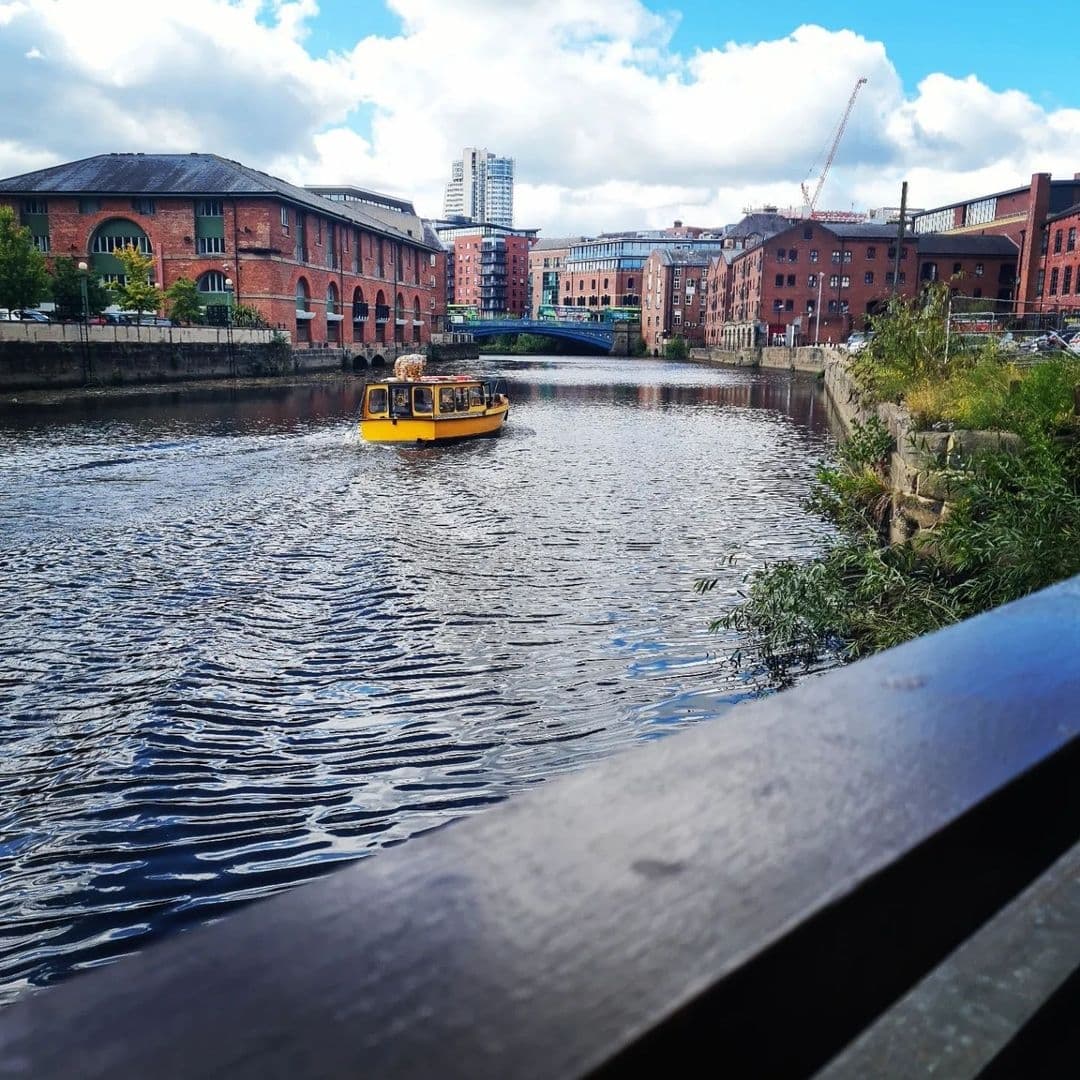 "Scenic view of a yellow water taxi on the river near Aire Bar with historic red brick buildings in the background and a bridge in the distance, reflecting the vibrant urban atmosphere."