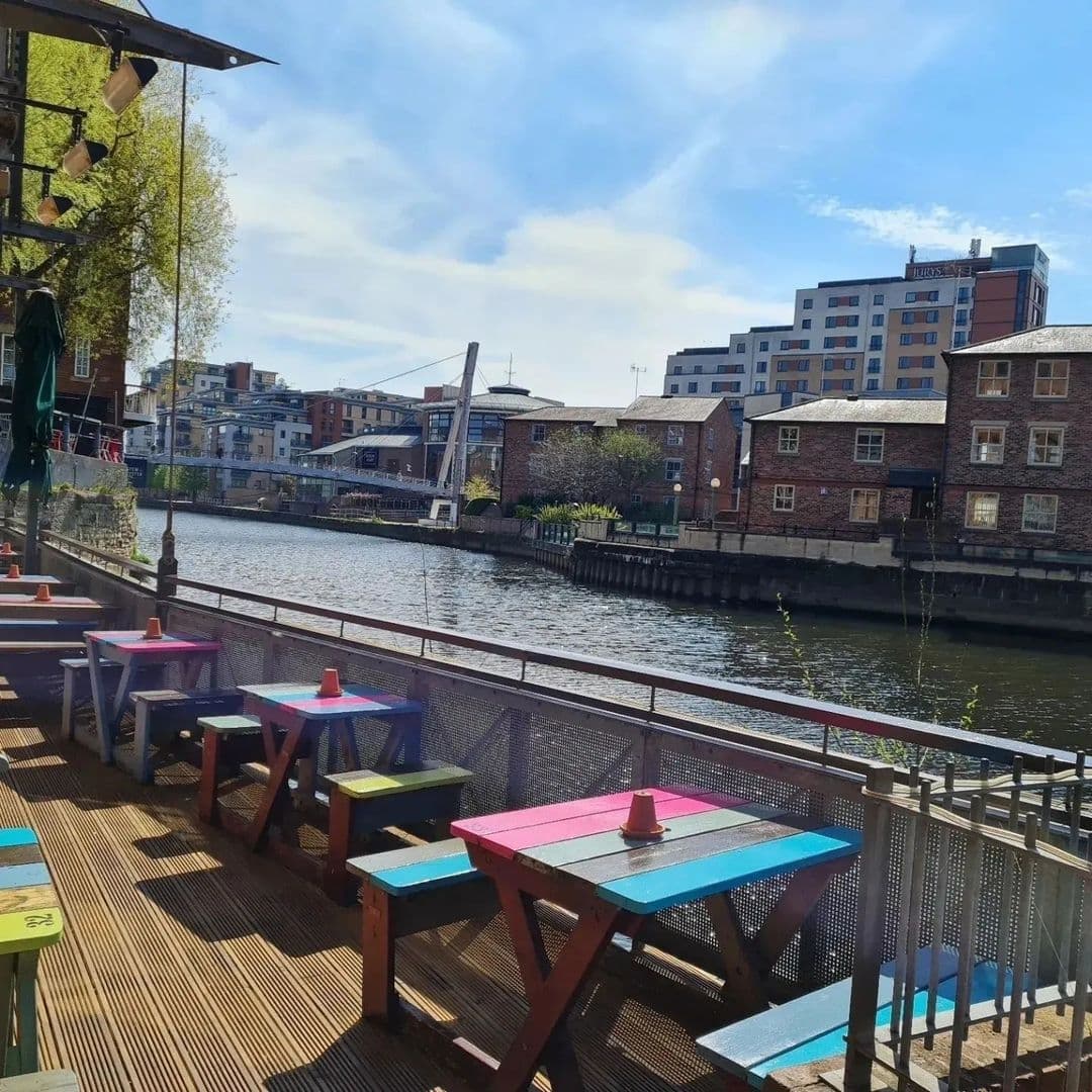 Outdoor seating at Aire Bar with colorful picnic tables overlooking a calm river, with a pedestrian bridge and urban buildings in the background.