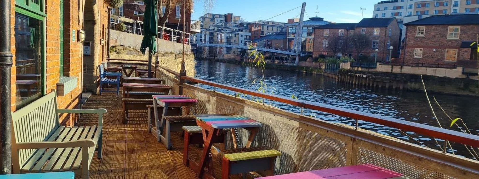 Outdoor seating area of Aire Bar with colorful tables overlooking a river, with urban buildings in the background.