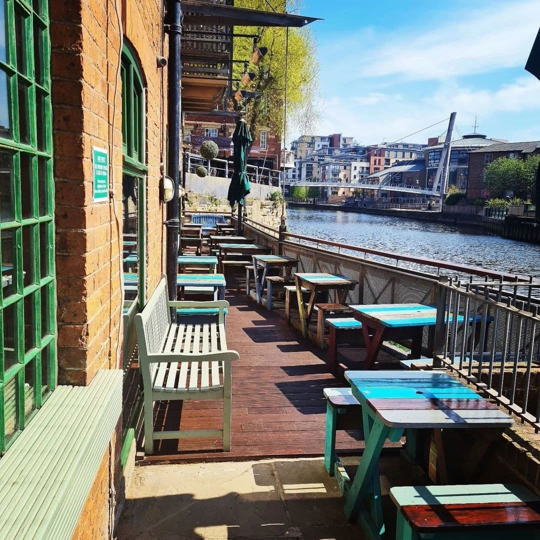 Sunny riverside outdoor seating at Aire Bar with wooden benches and tables overlooking the water and cityscape.