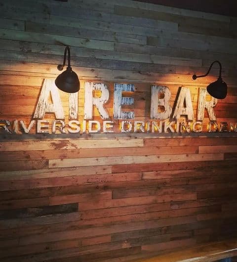 Illuminated sign of Aire Bar with the subtitle 'Riverside Drinking Den', mounted on a rustic wooden wall flanked by two hanging black lamps.