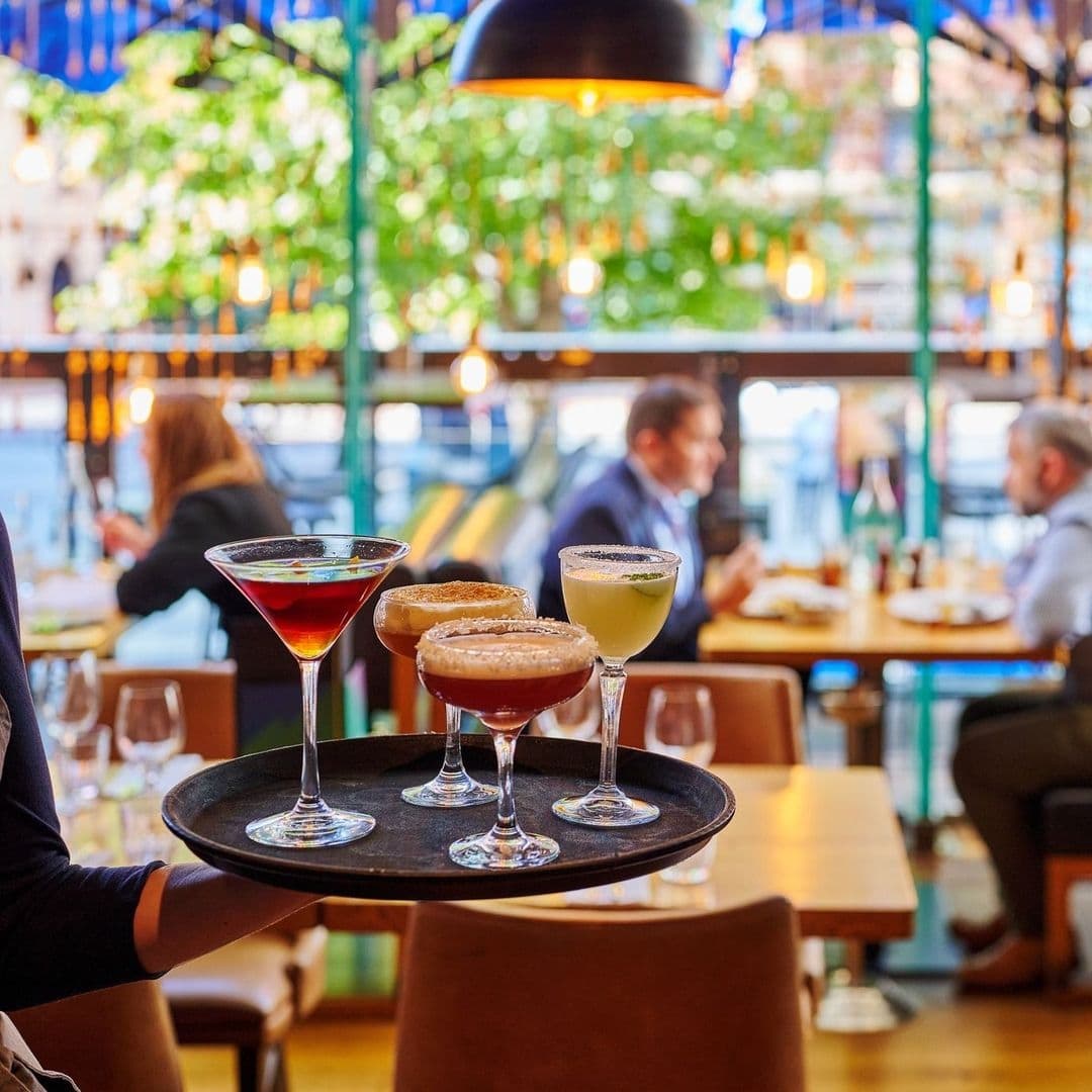 Alt text: "A server holding a tray with four stylish cocktails at Blackhouse, with patrons enjoying the vibrant atmosphere in the background."