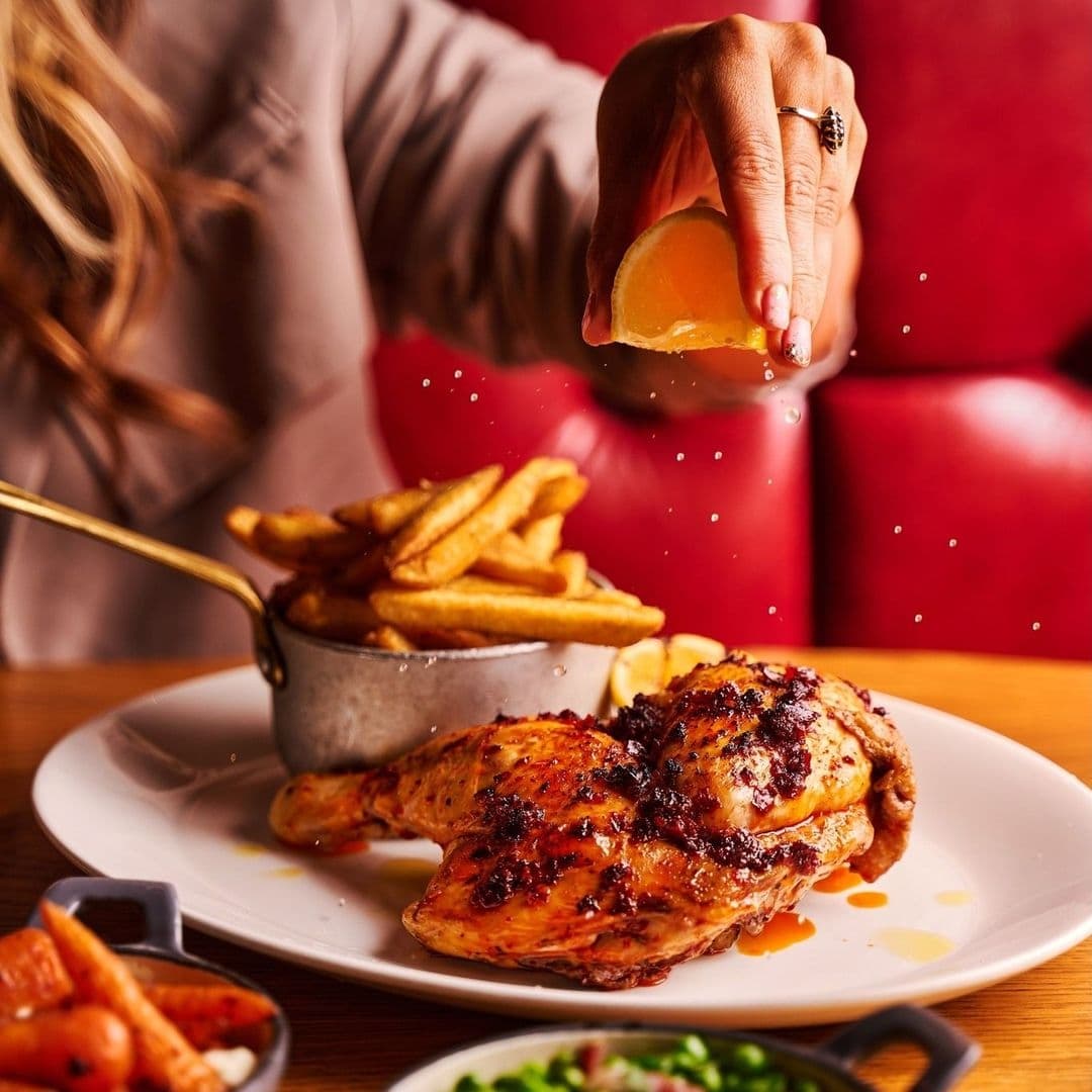 Person squeezing fresh lemon juice over a roasted chicken meal at Blackhouse, with golden fries and grilled vegetables on the side.