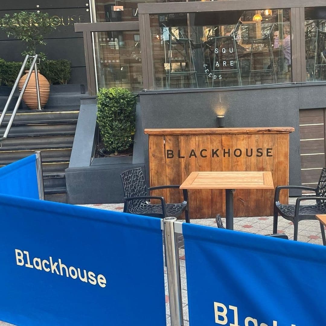 Outdoor seating area of Blackhouse with a wooden host stand prominently displaying the establishment's name, surrounded by blue barriers and black chairs, with a view of the restaurant's glass facade in the background.
