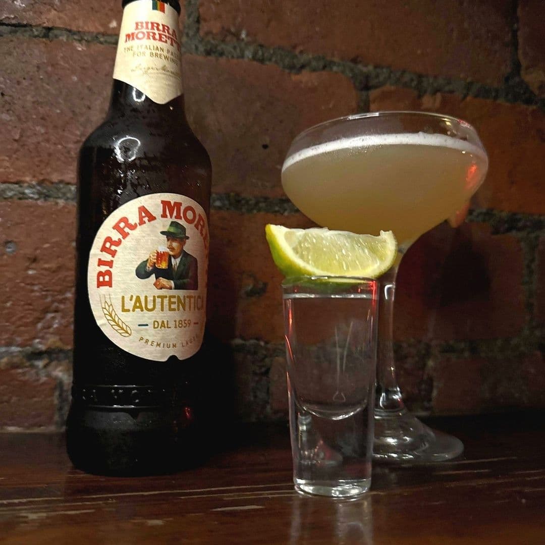 A chilled cocktail with a lime wedge on the rim and a bottle of Birra Moretti alongside a shot glass on a brick surface at Call Lane Social.