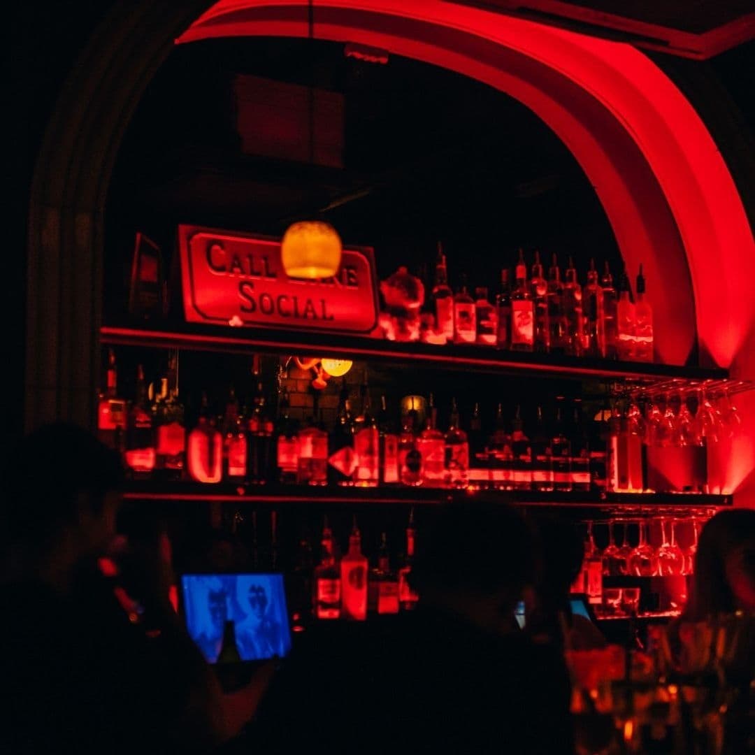 Interior of Call Lane Social with ambient red lighting showcasing the bar area and liquor bottles, with patrons in the foreground.