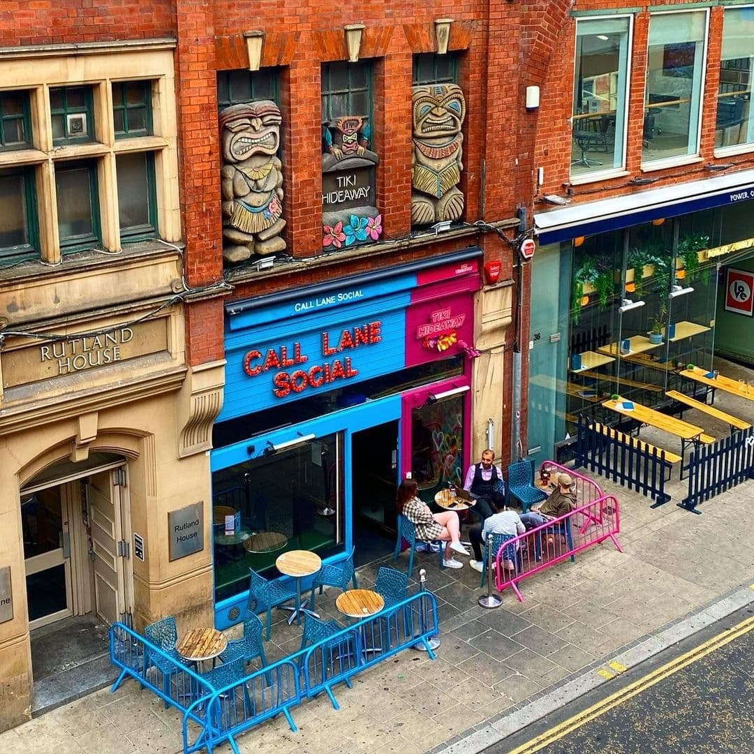 Exterior view of Call Lane Social with patrons sitting at colorful outdoor seating, vibrant blue and pink facade, and unique Tiki Hideaway decorations above the entrance.