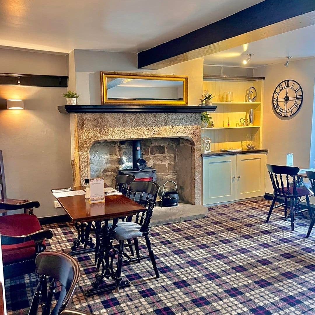 Cozy interior of Fox & Hounds featuring traditional dining furniture, a stone fireplace, tartan carpet, and classic decor with a large wall clock.