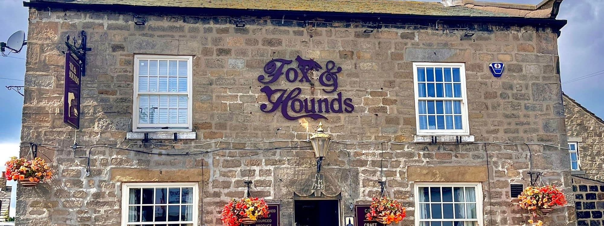 Exterior view of the Fox & Hounds, a traditional stone building with a purple sign, white-framed windows, and hanging flower baskets.