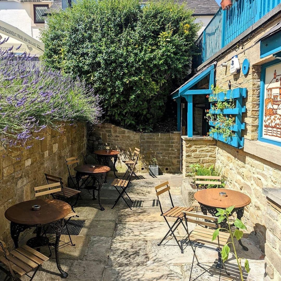Sunny outdoor seating area at Ilkley Moor Vaults with wooden tables and chairs, surrounded by lush greenery and flowering plants.